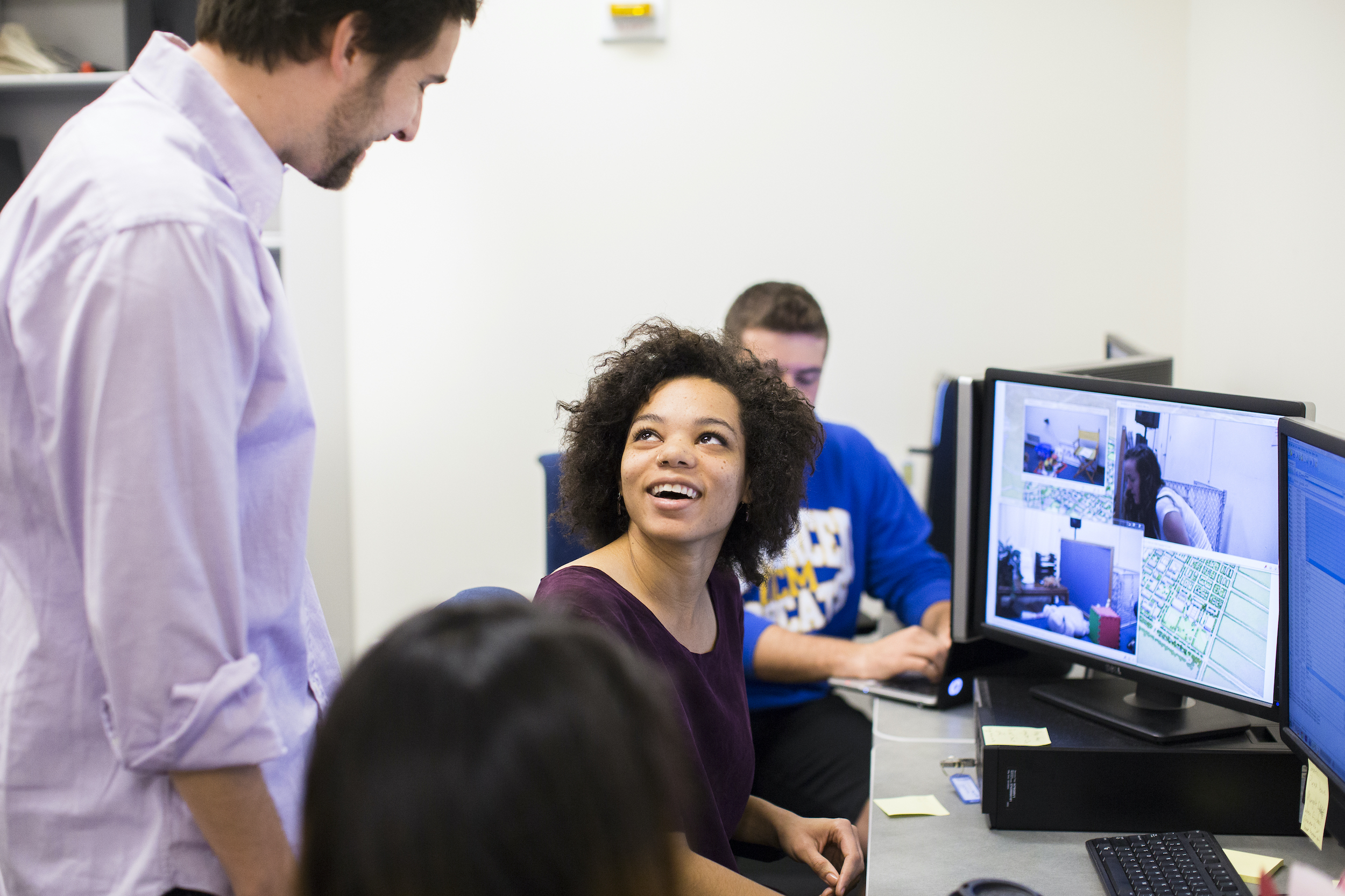 A young woman is sitting in front of a computer. She is looking back towards the camera and smiling. Several other students are working nearby.