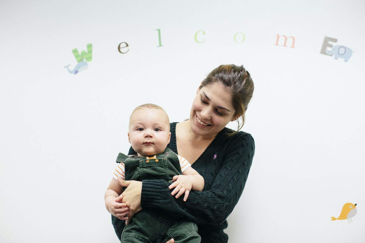 A young woman is holding a baby. The baby is looking into the camera. The word "Welcome" is on the wall behind them.