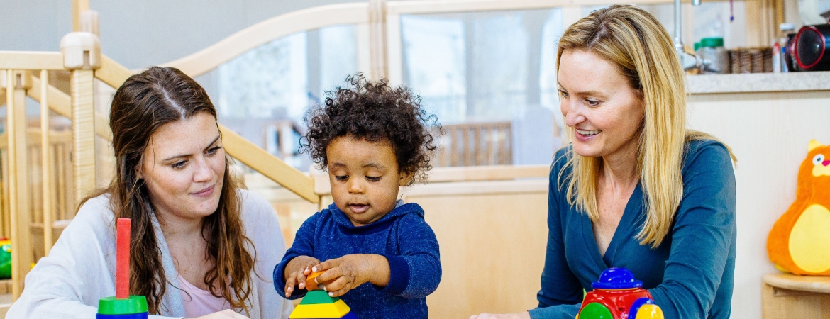 Two women smiling while watching an infant stack toy rings