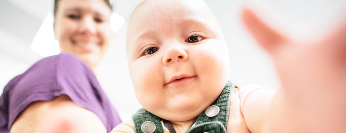 A young infant reaching towards the camera. A woman is smiling behind him.